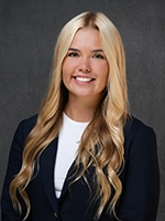 Professional headshot of a smiling blonde woman with long wavy hair, wearing a dark blazer over a white top against a gray background.