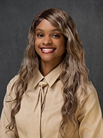 Studio headshot of a smiling person with long wavy hair, wearing a beige button-up shirt against a dark gray background.