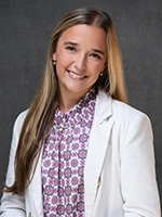 Smiling woman with long blonde hair wearing a white blazer over a patterned blouse, posed against a gray studio backdrop.