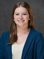 Professional headshot of a smiling woman with long brown hair, wearing a blue blazer over a light top against a gray background.