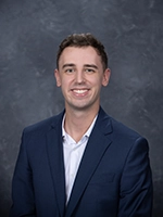 Professional headshot of a man wearing a navy blazer and white shirt, smiling against a gray background.