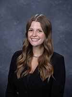 Professional headshot of a young woman with long brown hair, wearing a black blazer and smiling.