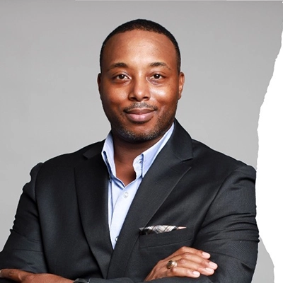 Professional headshot of a man in a black suit with a light blue shirt, smiling confidently with arms crossed.