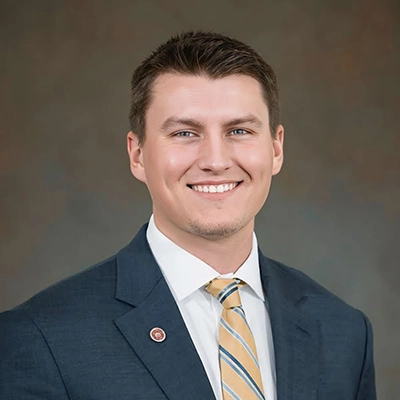 Professional headshot of a young man in a suit and tie, smiling confidently.