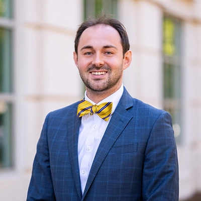 Professional portrait of a man in a blue suit and yellow bow tie, smiling in front of a building.