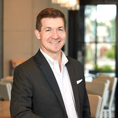 Professional portrait of a smiling man in a suit, standing in a modern interior setting.
