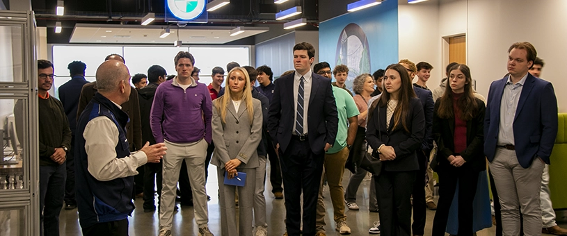 A group of individuals listening attentively to a woman speaking in an automotive workshop setting, with various cars and equipment visible in the background.