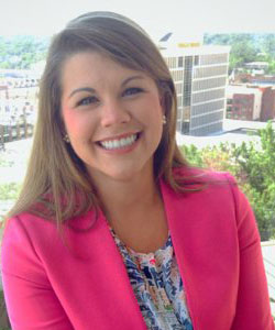 A smiling woman wearing a pink blazer, posing outdoors with a city skyline in the background.