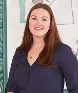 Professional headshot of a woman with long brown hair, wearing a navy blouse, smiling against a light, artistic background.