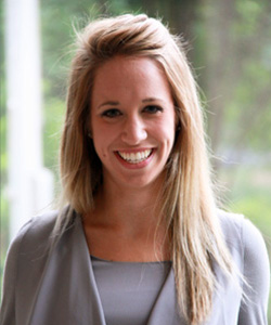 Smiling woman with long blonde hair wearing a grey top standing outdoors.