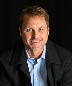 Headshot of a smiling man with blue eyes, wearing a black jacket over a light blue shirt, against a dark background.