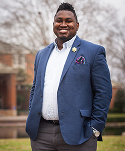 A smiling man in a blue blazer with a patterned pocket square, standing outdoors.