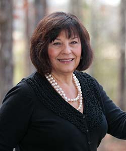 A smiling middle-aged woman wearing a black blouse and a pearl necklace, standing outdoors among trees.