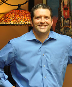 Smiling man in a blue shirt standing confidently with a decorative lamp in the background.