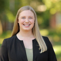 Smiling woman in a professional blazer standing outdoors with a blurred background of greenery.