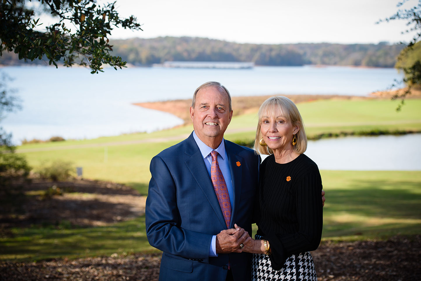 Older man and his wife holding hands and standing together with grass and lake in the background.