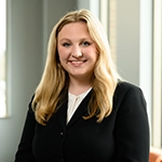 Smiling person with long blonde hair wearing a black top, standing indoors near a window.