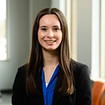 Portrait of a smiling woman in a black blazer and blue blouse in an office setting.