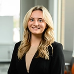 Smiling blonde woman in a black blazer standing indoors with a softly blurred background.