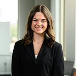 Smiling person with shoulder-length dark hair wearing a black blazer, standing indoors near a window.
