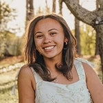 Smiling woman outdoors in a sunlit park with trees in the background.