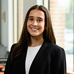 Smiling woman in a black blazer standing indoors near a window.