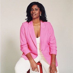 Studio portrait of a woman wearing a bright pink blazer and white pants, seated on a stool against a light background.