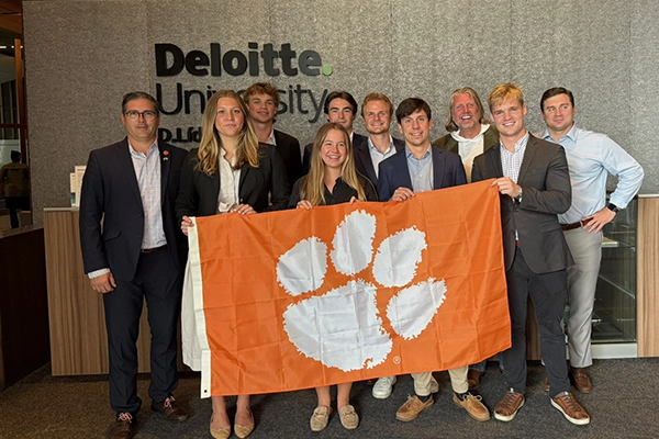 Group of people posing indoors in front of a Deloitte University sign while holding an orange Clemson Tigers flag with a white paw print.