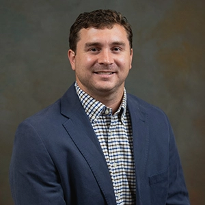 Smiling man in a blue blazer and checkered shirt against a neutral studio background.