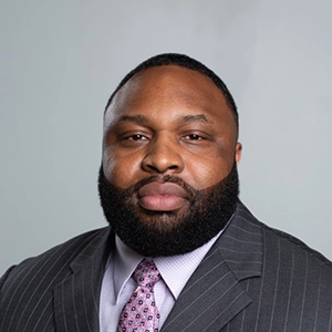 Headshot of a man with a full beard wearing a dark pinstripe suit, light shirt, and patterned tie against a gray background.