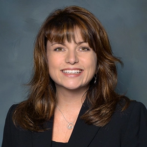 Professional headshot of a smiling woman with shoulder-length brown hair, wearing a dark blazer against a gray studio background.