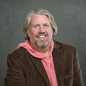 Smiling middle-aged man with gray hair and a beard, wearing a brown blazer over a pink hoodie, posed against a gray studio backdrop.