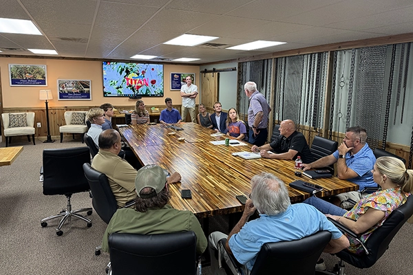 Group of people seated around a large conference table in a meeting room, listening to a man standing and speaking.