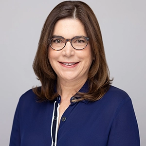 Studio headshot of a smiling woman with shoulder-length brown hair, wearing glasses and a navy blouse against a light gray background.