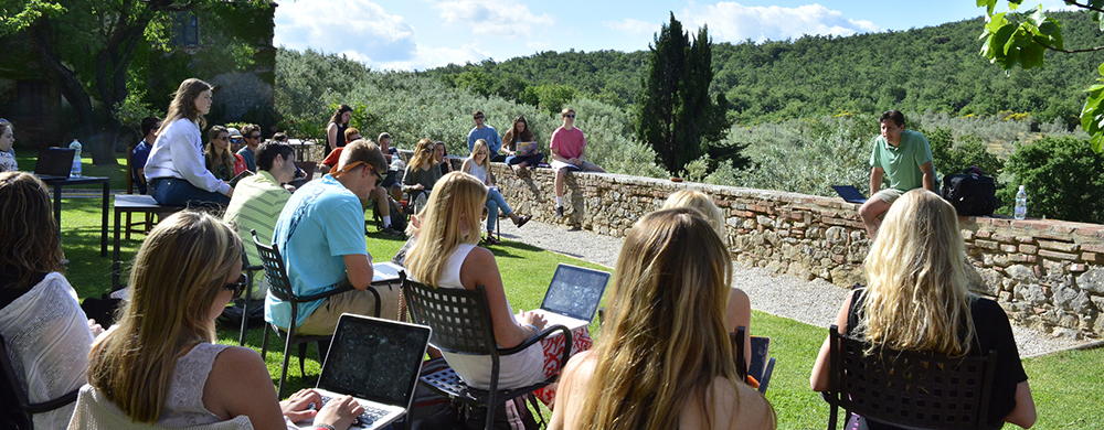 A group of people sitting outdoors in a scenic landscape, engaged in a discussion or a workshop, with laptops open in front of them.