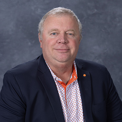 Professional headshot of a man in a dark blazer and patterned shirt featuring an orange and white color scheme.