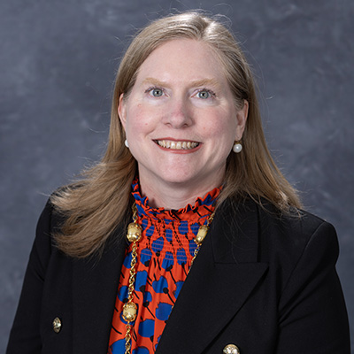 A professional headshot of a woman wearing a colorful patterned blouse and a black blazer, smiling at the camera.