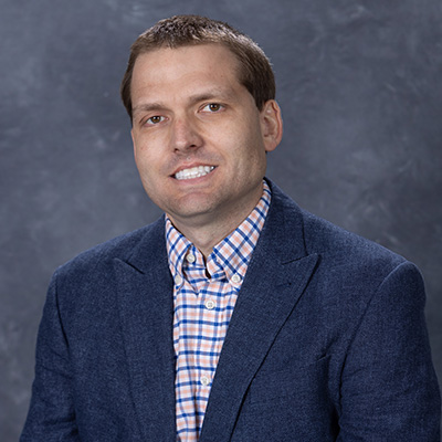 Professional portrait of a man in a blue blazer and plaid shirt, smiling against a gray background.