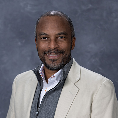 Professional headshot of a smiling man in a light blazer and gray sweater.