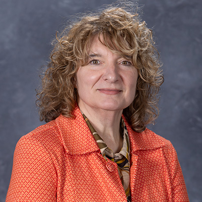 Headshot of a woman with curly hair, wearing an orange blazer and a patterned blouse, set against a gray background.