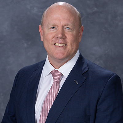 Professional headshot of a man in a dark suit and pink tie, smiling against a gray background.