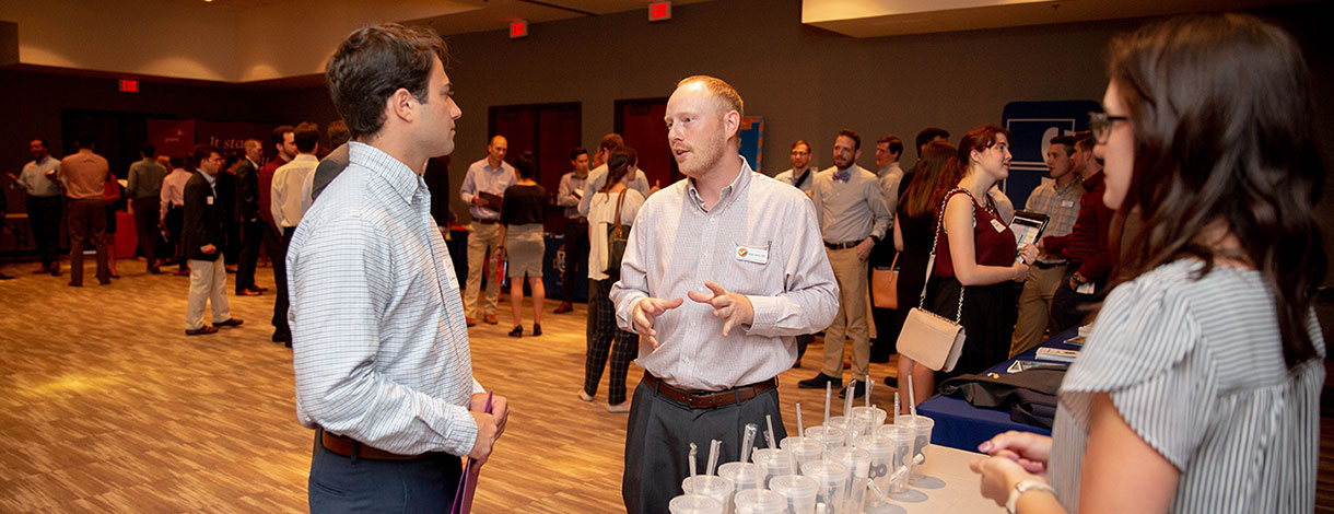 A networking event with attendees engaged in conversation, showcasing a professional atmosphere and various informational booths in the background.