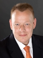 Professional headshot of a man wearing a dark suit and orange tie, with short hair and a friendly expression.