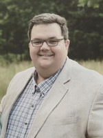 Smiling man in a light blazer with a plaid shirt, standing outdoors in a natural setting.