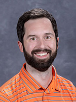 A professional headshot of a smiling man with a beard, wearing an orange striped polo shirt.