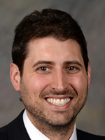Formal headshot of a smiling man with dark hair and a beard, wearing a suit and tie.