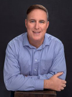 Professional headshot of a smiling man with short hair, wearing a light blue shirt, leaning slightly forward with arms crossed.