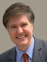 Professional headshot of a man with short, light brown hair, smiling confidently in a suit and tie.