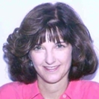 Professional headshot of a woman with curly hair, wearing a pink blouse and smiling.