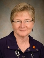Professional headshot of a woman with short blonde hair and glasses, wearing a purple shirt with a Clemson University logo.